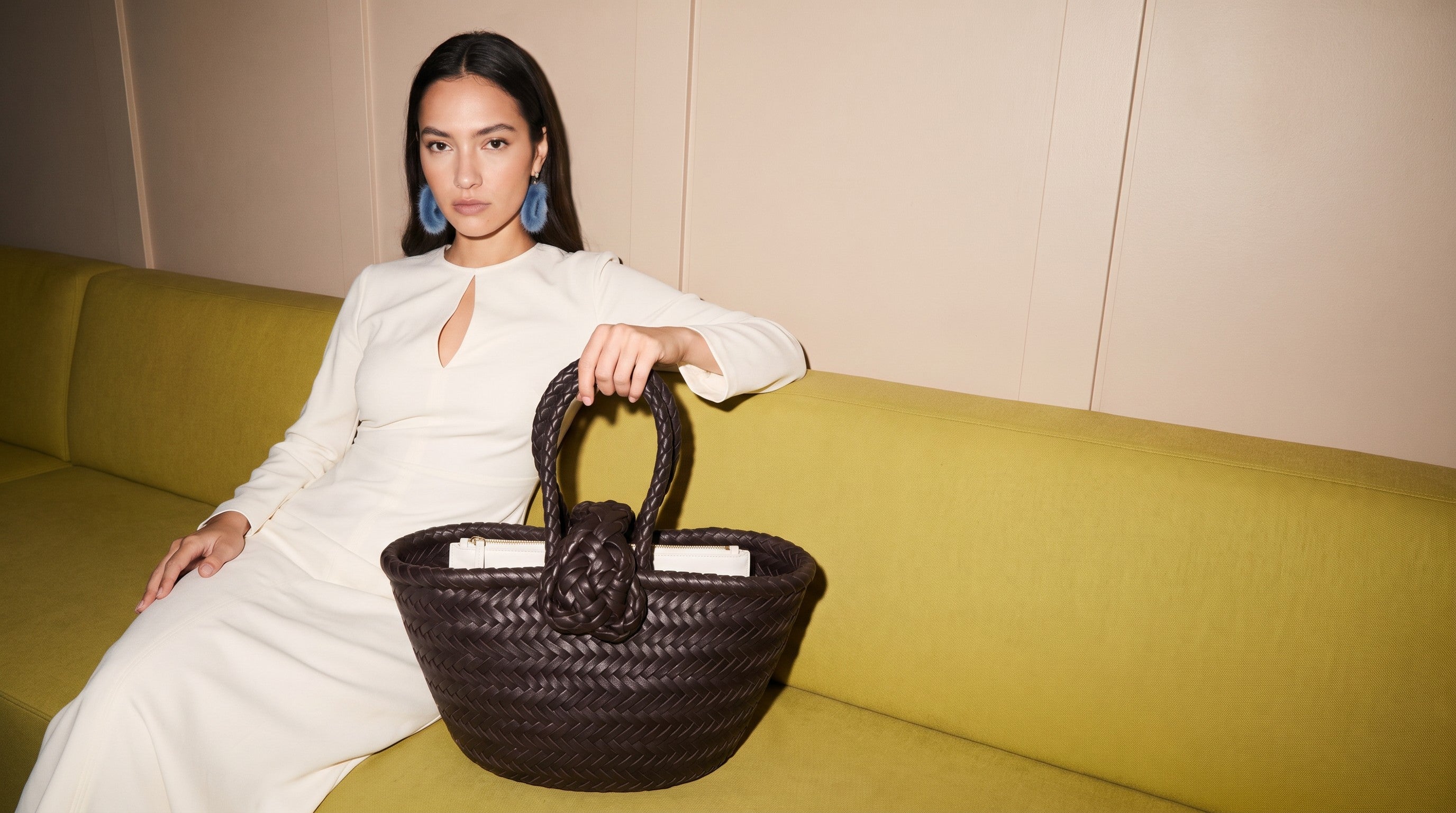 Femme en robe crème assise sur un canapé olive, tenant à la main un sac cabas en cuir tressé marron dans une ambiance méditerranéenne chaude